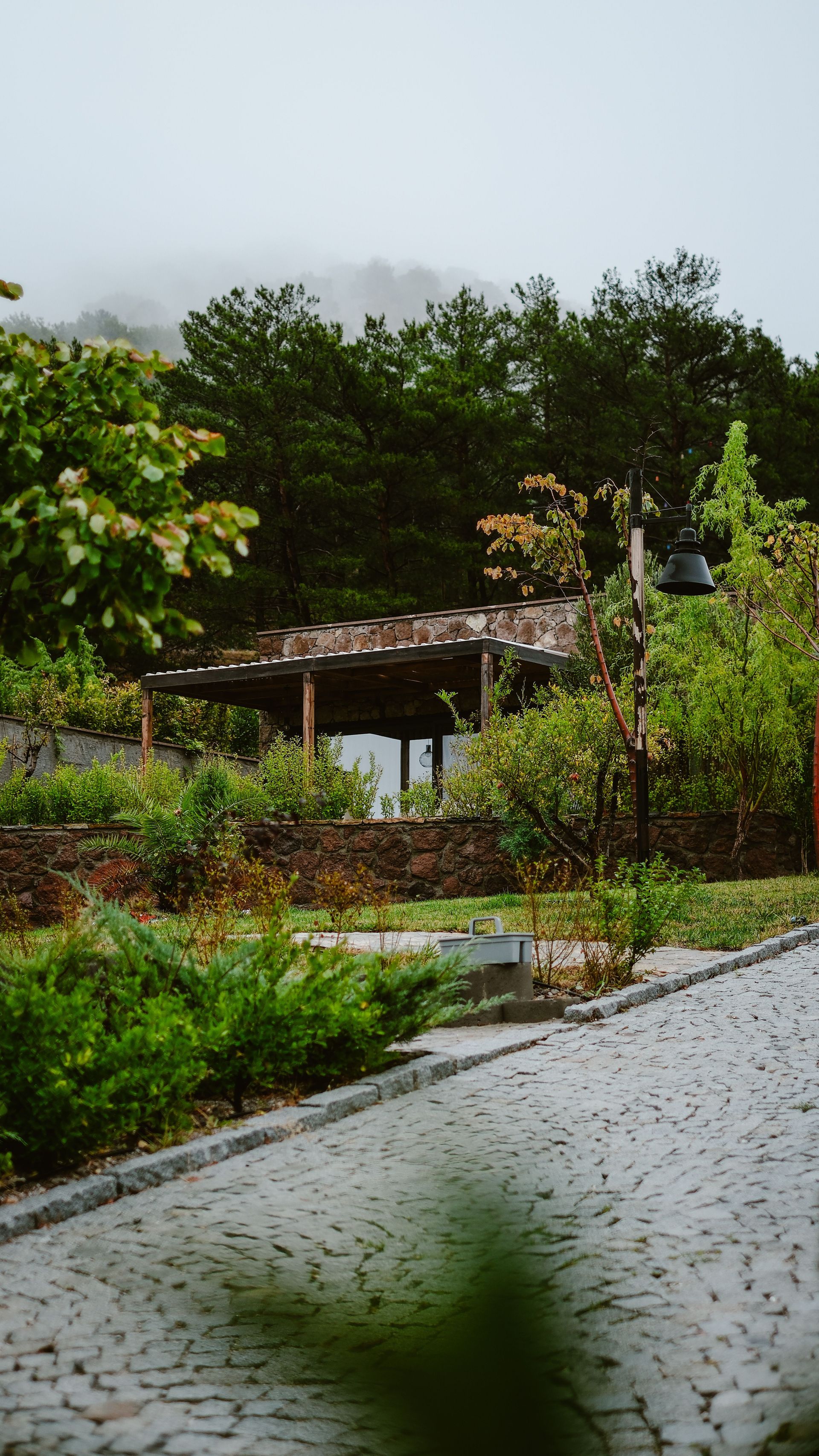 A cobblestone path leads to a rustic pavilion surrounded by lush greenery and trees, creating a tranquil outdoor setting.