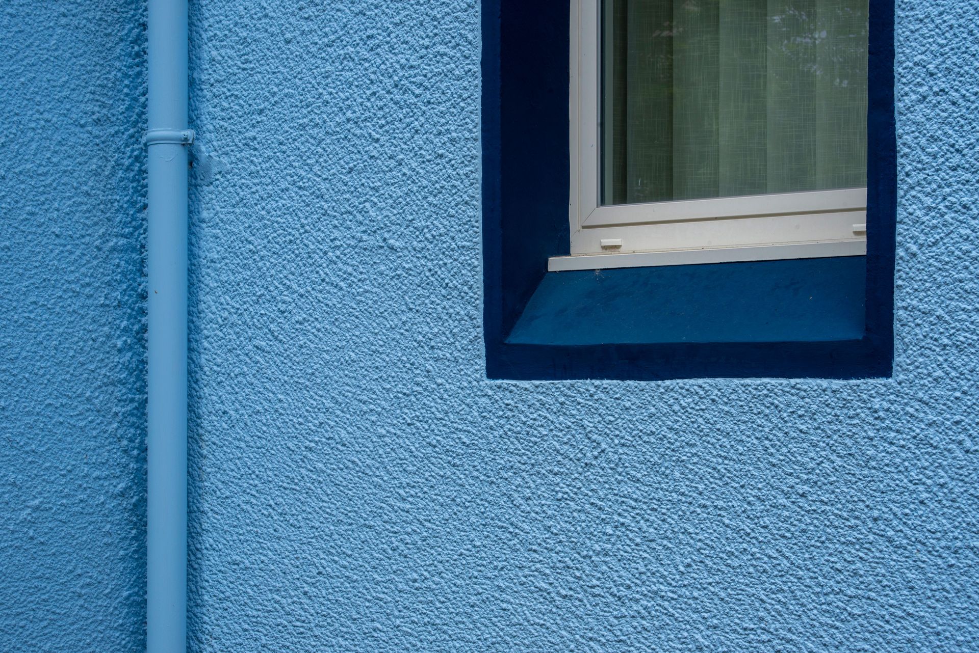 Close-up of a blue textured wall featuring a window and a vertical drainpipe.