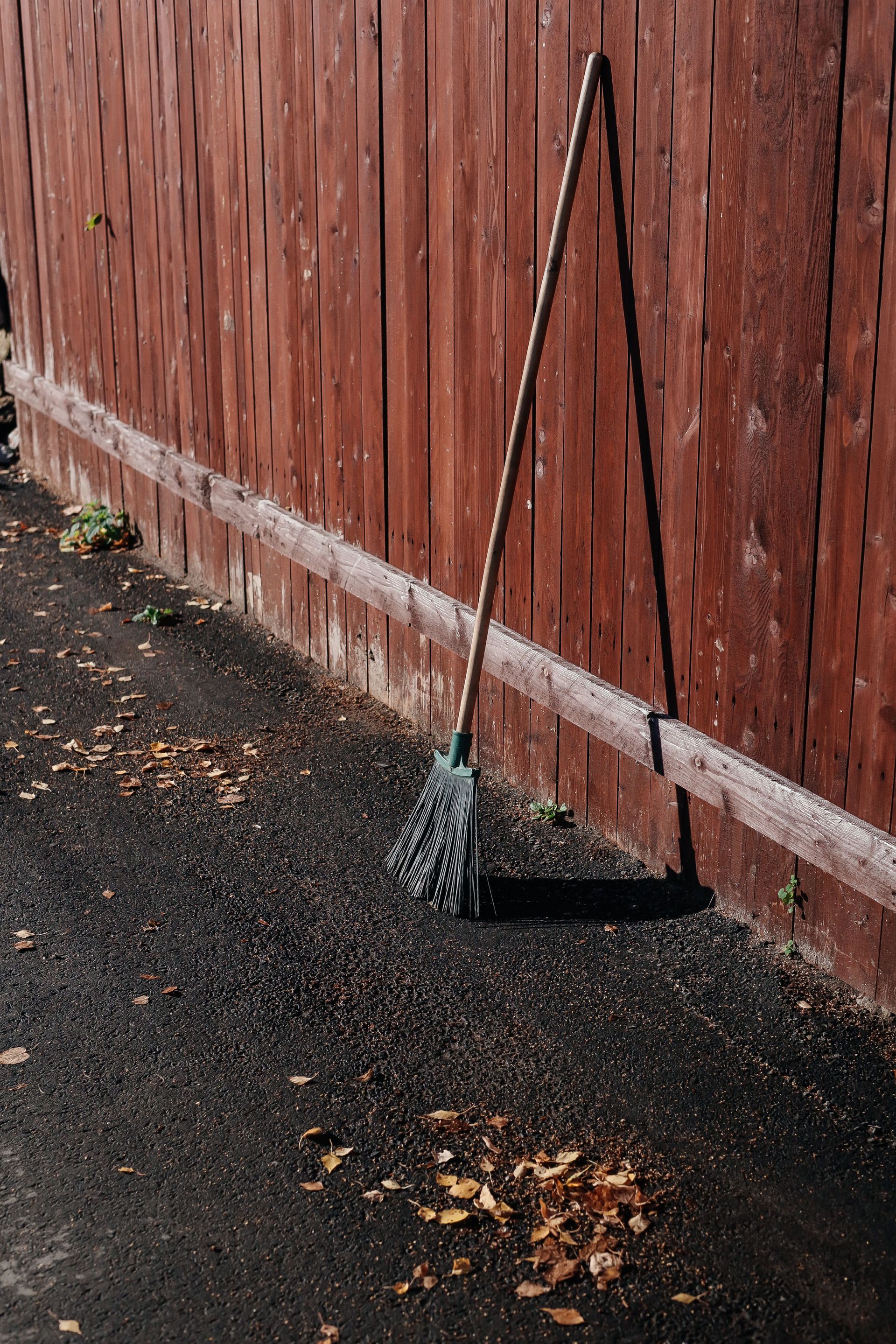 A solitary broom leaning against a red wooden fence on a sunny autumn day.